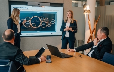 2 Frauen halten eine Präsentation in einem Konferenzraum. Am Tisch sitzen zwei Männer mit ihrem Notebook